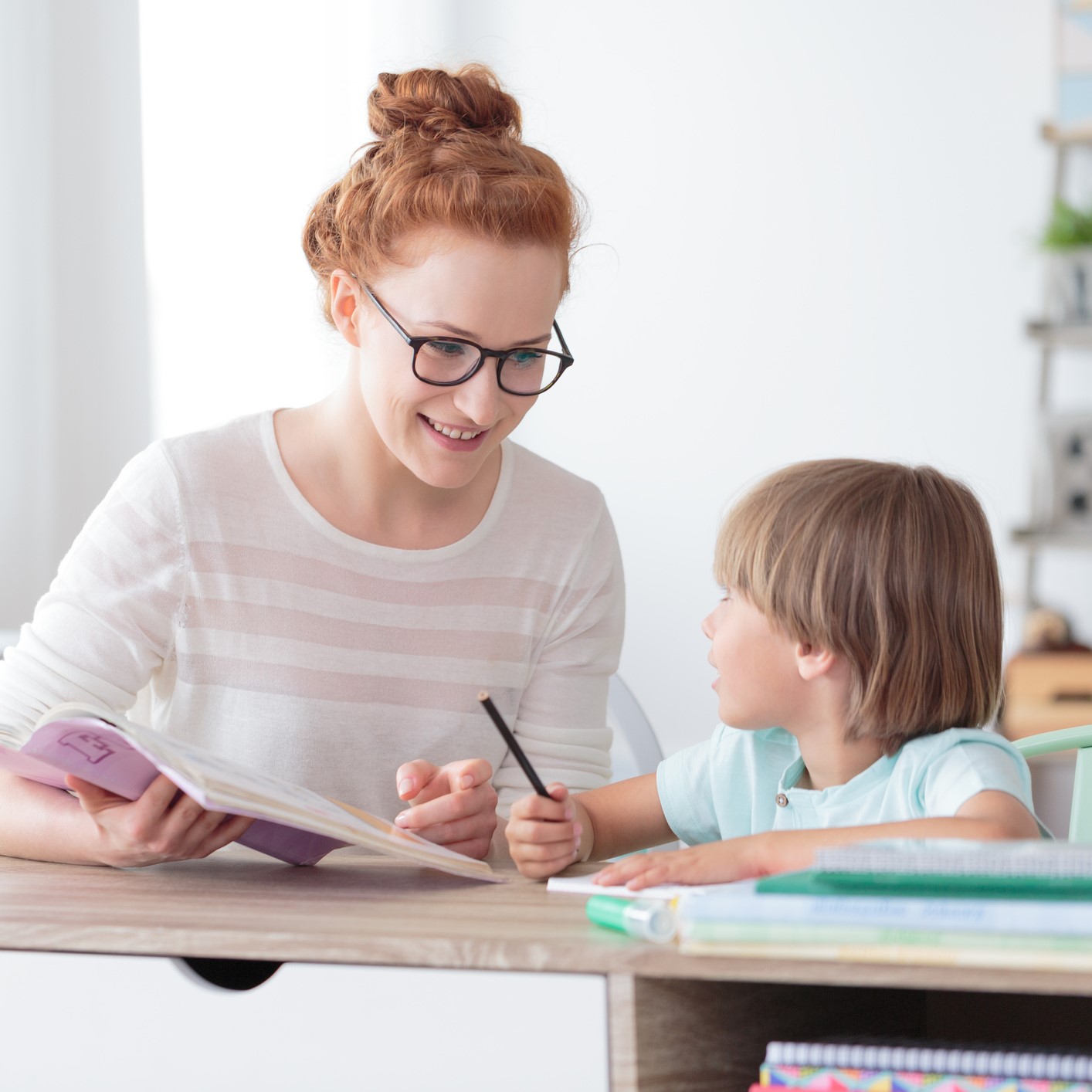 Smiling mother helping son with homework while sitting at desk with notebooks in home office